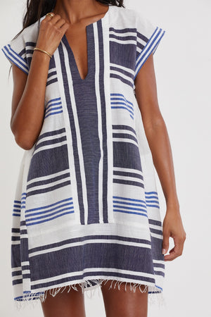 A person wearing the Lemlem Elina Caftan, a short-sleeved, V-neck dress featuring blue, navy, and white stripes with a fringed hem and traditional tibeb weaving inspired by hand-woven Ethiopian designs.