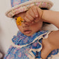 A child wearing a floral outfit and a Louise Misha Baby Lajik Sun Hat made of cotton voile holds a small yellow flower up to their face, partially covering one eye.