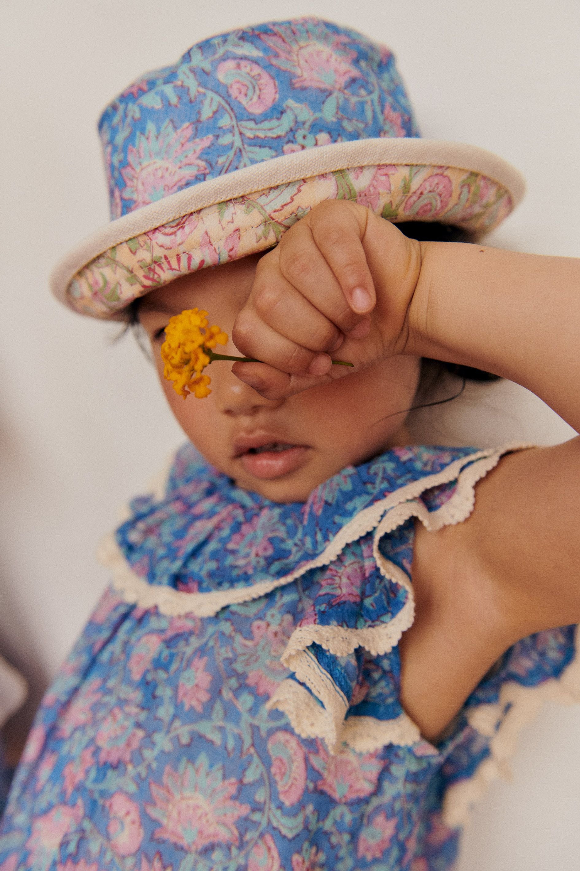 A child wearing a floral outfit and a Louise Misha Baby Lajik Sun Hat made of cotton voile holds a small yellow flower up to their face, partially covering one eye.