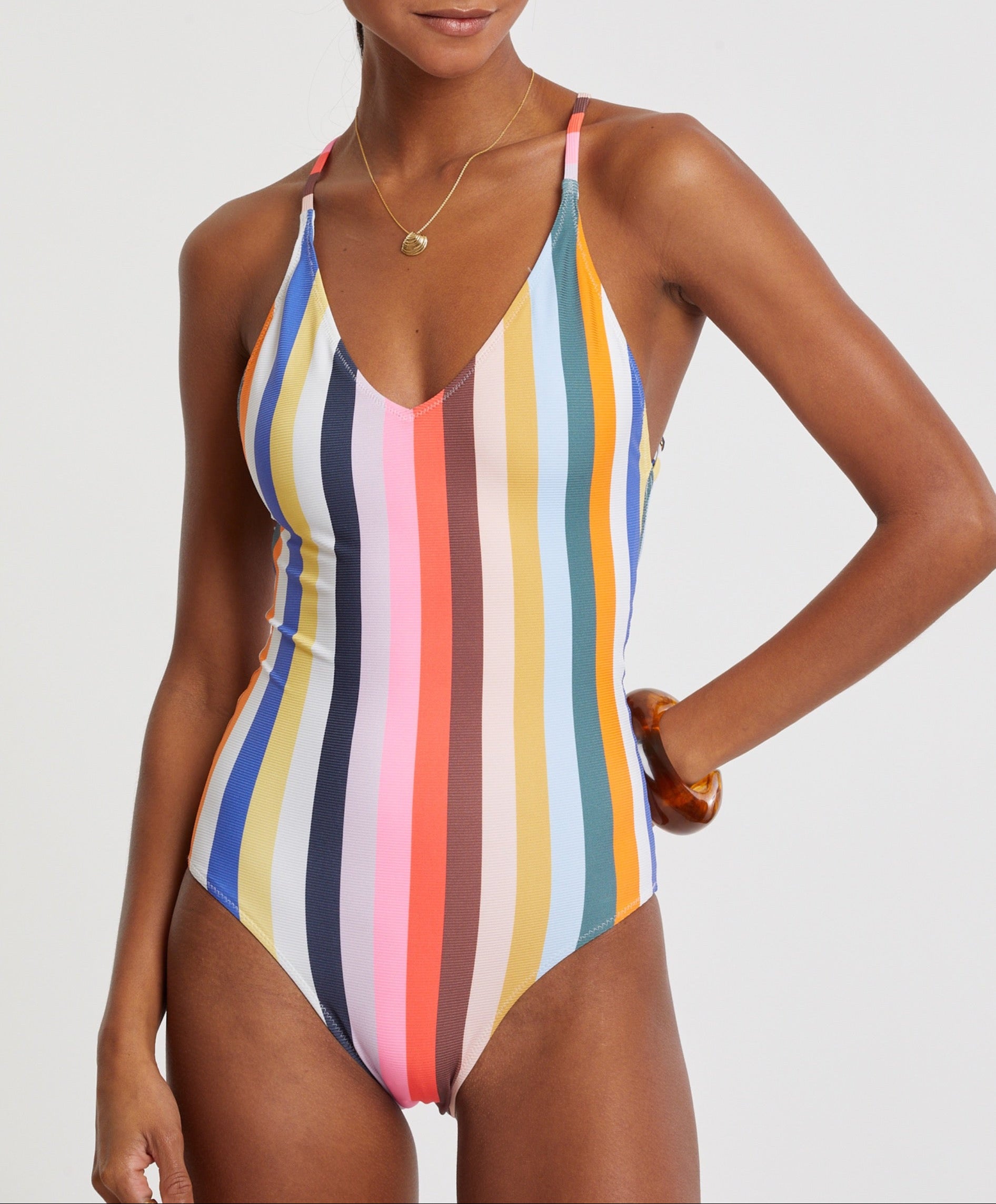 A woman wears the Lemlem Retiro One Piece, featuring vertical multicolored stripes, thin shoulder straps, and medium-coverage legs, accessorized with a necklace and bracelet.