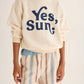 A person stands against a plain background wearing a cream cotton sweatshirt with “Yes, Sun;” printed in blue and Cozmo Boys' Zion Bermuda Shorts featuring stripes and an elastic waistband.