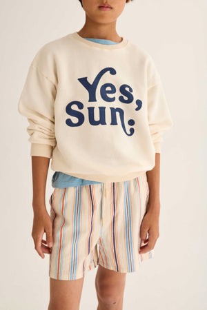 A person stands against a plain background wearing a cream cotton sweatshirt with “Yes, Sun;” printed in blue and Cozmo Boys' Zion Bermuda Shorts featuring stripes and an elastic waistband.