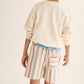A child stands facing away, wearing a cream sweatshirt paired with Cozmo Boys' Zion Bermuda Shorts, which feature a striped design, cotton fabric, an elastic waistband, and a back pocket.
