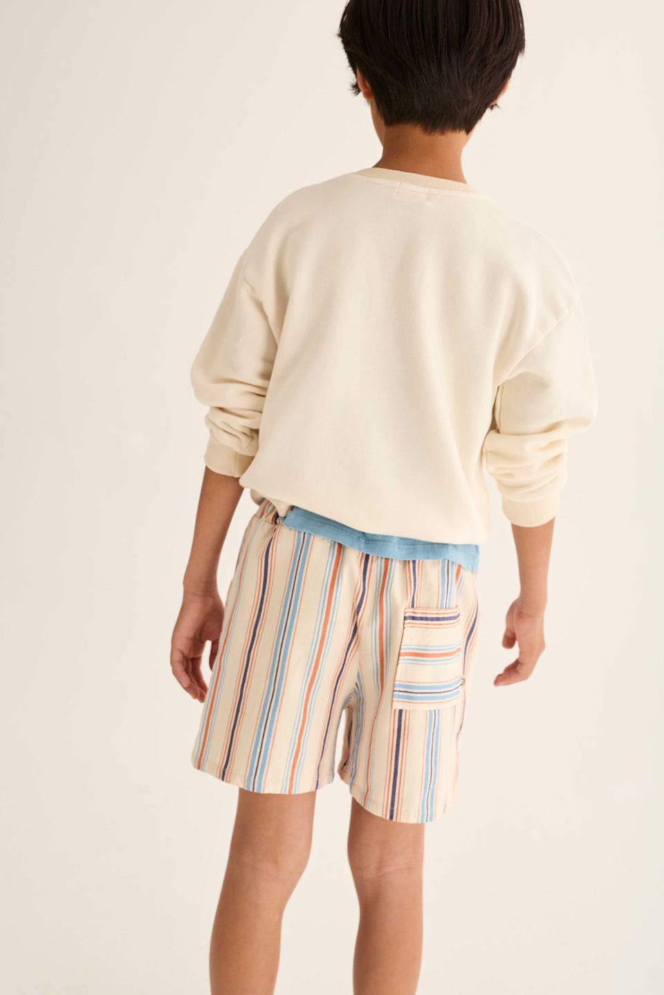 A child stands facing away, wearing a cream sweatshirt paired with Cozmo Boys' Zion Bermuda Shorts, which feature a striped design, cotton fabric, an elastic waistband, and a back pocket.