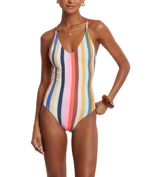 A woman models the Lemlem Retiro One Piece, featuring vertical multicolored stripes and an adjustable cross-back design, standing with one hand on her hip against a plain white background.