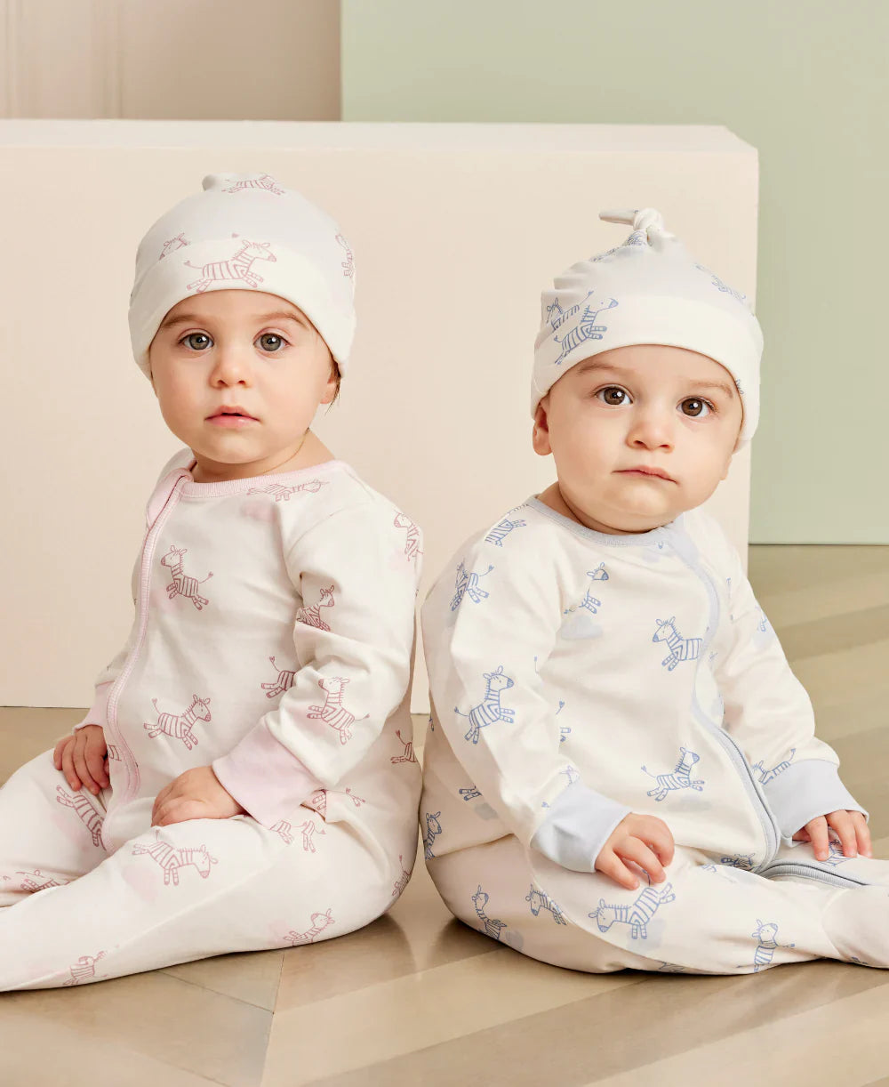 Two babies dressed in matching Kissy Kissy Love Dreamy Zebras Zip Footie outfits, complete with cozy hats, sit together on the floor. Crafted from soft Pima cotton, one outfit is pink and the other is blue, each featuring handy mitten-cuffs to keep little hands warm.