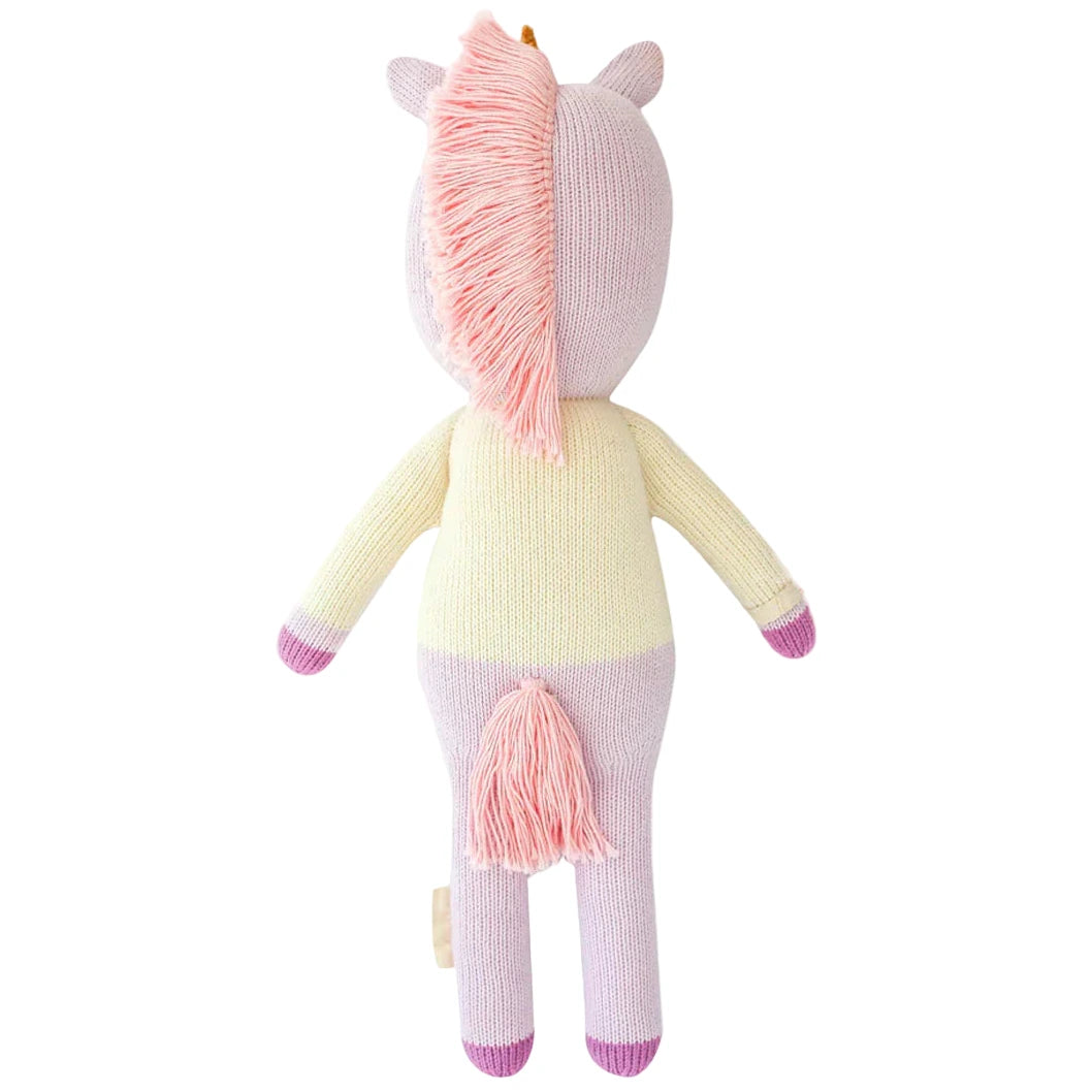Zoe the Unicorn - Regular 20" by Cuddle + Kind is a magical knitted plush with a pink mane and tail, cream-colored body, and purple hooves, pictured from behind as if journeying into a mystical forest.