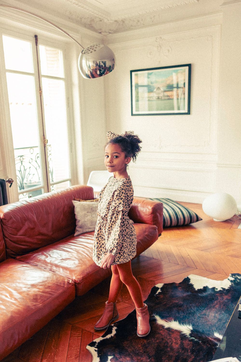 A young girl wears the Louis Louise Girls' Umyma Corduroy Leopard Dress with ruffles and boots, standing on a cowhide rug in a chic living room featuring a brown leather sofa and large windows.