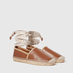 The Gaia Whipstitched Flat by Stella McCartney features brown leather, woven soles, and white ankle ties with logo accents, showcasing natural textures and a handcrafted finish for an elevated flat sandal style.
