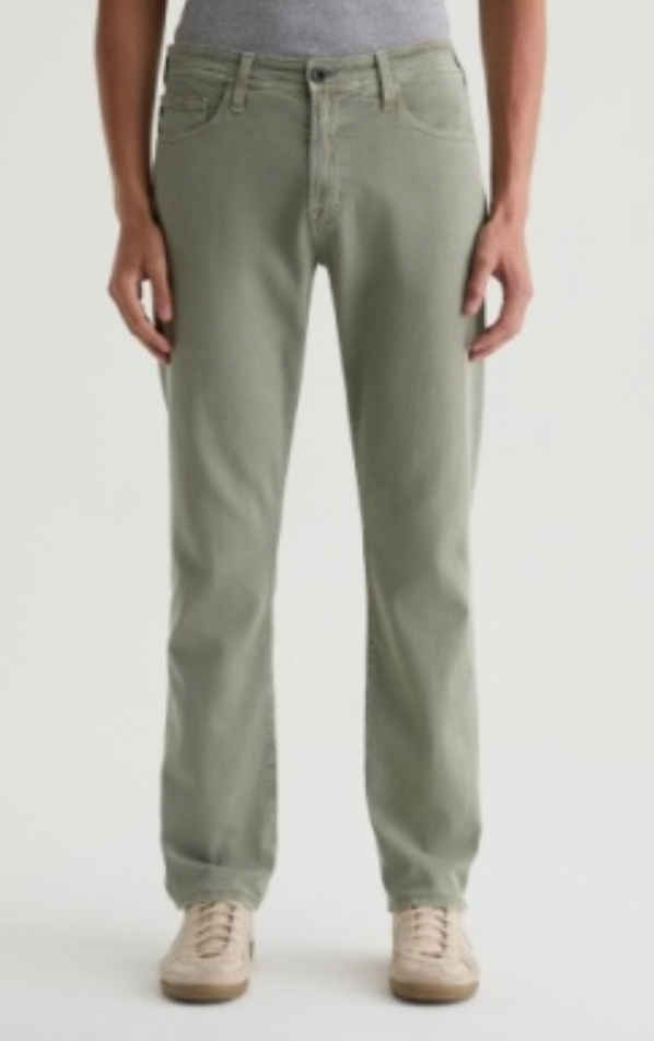 A man wearing AG Jeans’ Everett Slim Straight jeans in sage green, featuring a mid-rise waist and classic five-pocket design, paired with beige sneakers, against a white background.