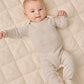 A baby wearing the Quincy Mae Baby Footie in beige and white stripes with fold-over hand mitts lies on a quilted cream-colored surface, looking up with both arms raised slightly.