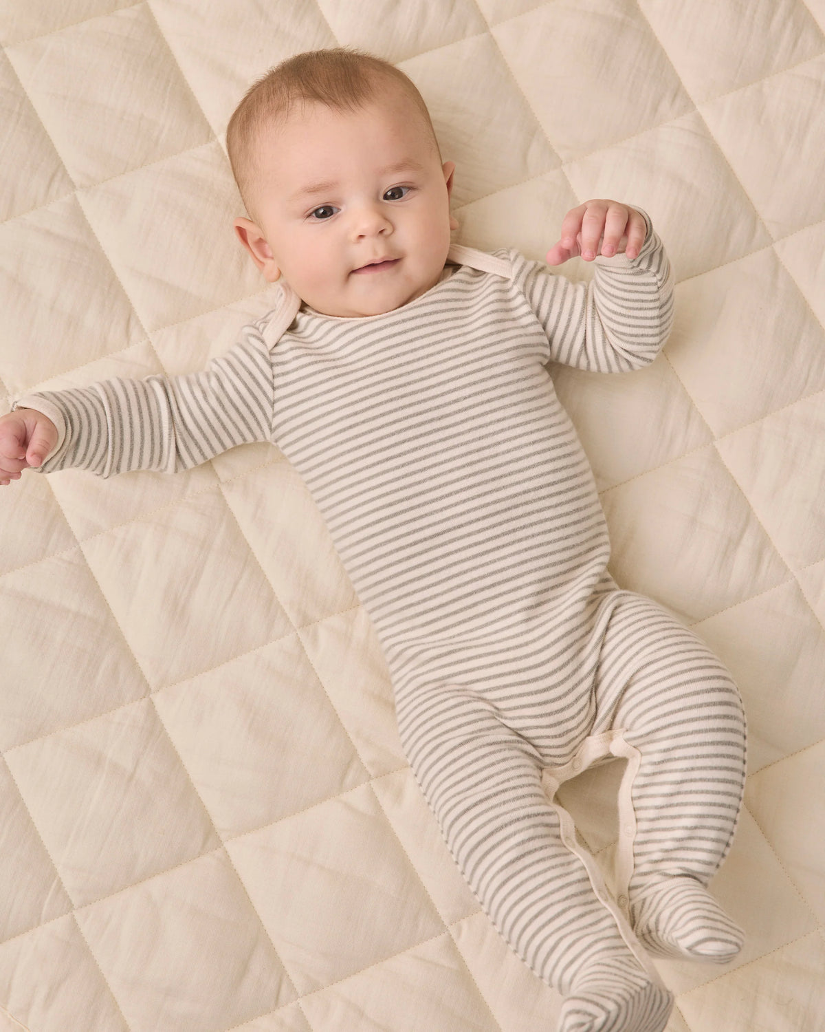 A baby wearing the Quincy Mae Baby Footie in beige and white stripes with fold-over hand mitts lies on a quilted cream-colored surface, looking up with both arms raised slightly.