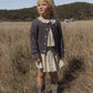 A young child stands in a grassy field wearing the Rylee & Cru Girls’ Smocked V Mini Skirt paired with a dark cardigan and gray cowboy boots, with rolling hills and a small building visible in the background.