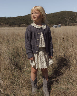 A young child stands in a grassy field wearing the Rylee & Cru Girls’ Smocked V Mini Skirt paired with a dark cardigan and gray cowboy boots, with rolling hills and a small building visible in the background.
