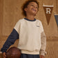 A smiling child stands indoors near wooden paneling and pennant banners, holding a vintage brown basketball and wearing the Boys' Relaxed Sweatshirt by Rylee & Cru for a classic athletic vibe.