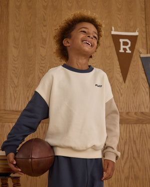 A smiling child stands indoors near wooden paneling and pennant banners, holding a vintage brown basketball and wearing the Boys' Relaxed Sweatshirt by Rylee & Cru for a classic athletic vibe.