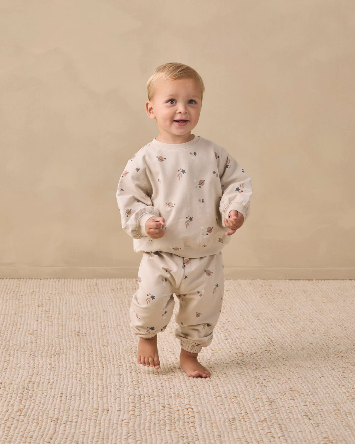 A toddler stands barefoot on a textured beige rug, wearing a matching cream-colored sweatshirt and Quincy Mae Baby Boys’ and Boys’ Oversized Sweatpants featuring a small floral print and an elastic waistband, against a plain beige background.