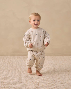 A toddler stands barefoot on a textured beige rug, wearing a matching cream-colored sweatshirt and Quincy Mae Baby Boys’ and Boys’ Oversized Sweatpants featuring a small floral print and an elastic waistband, against a plain beige background.