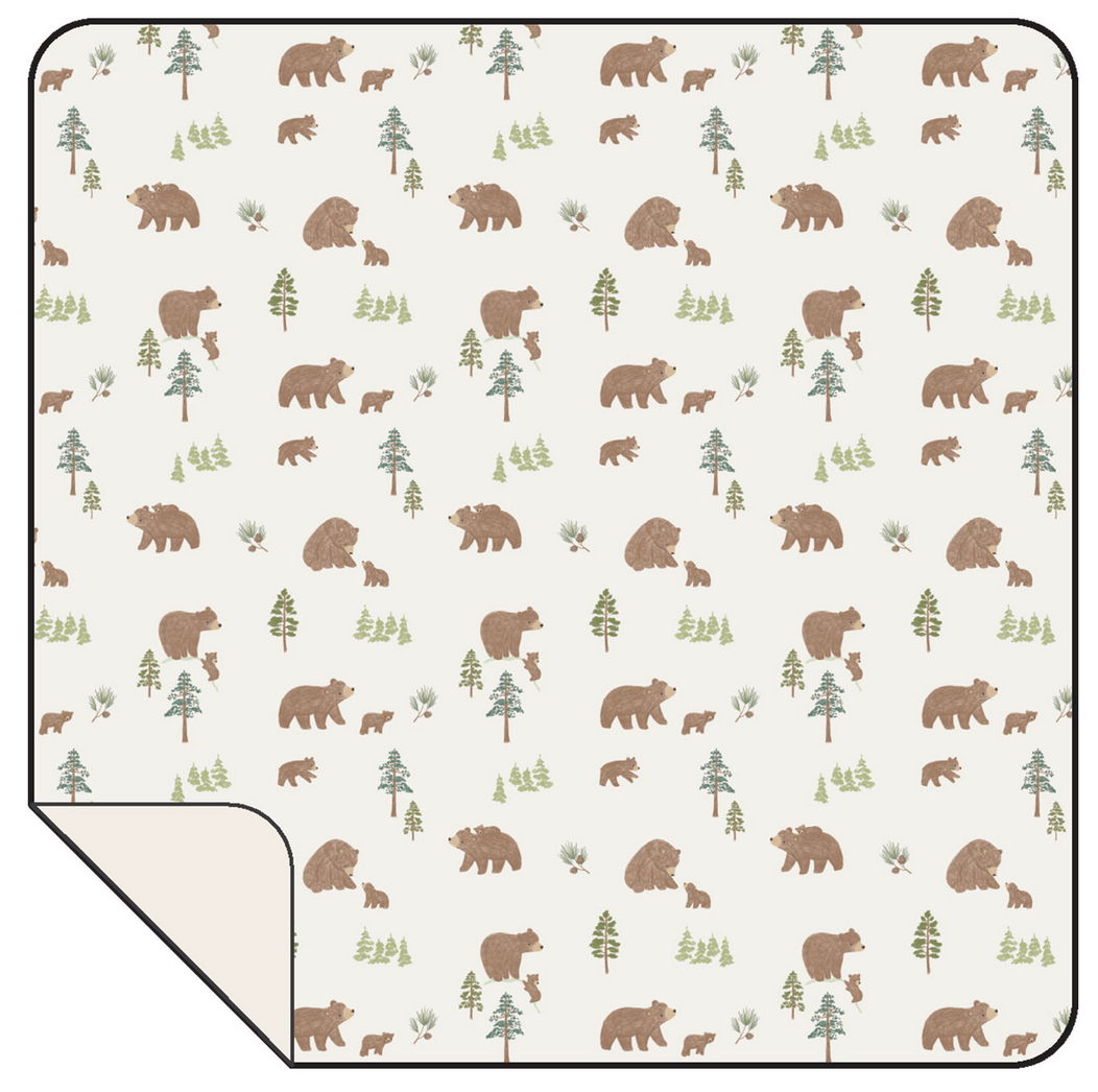 The Angel Dear Swaddle Blanket features a cream background with brown bear and green tree patterns, rounded corners, and a visible soft underside.