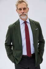 An older man with gray hair and a beard stands with his hands in his pockets against a plain light background, wearing the Sid Mashburn Ghost Blazer in green Italian hopsack, paired with a light blue shirt, a patterned red tie, and dark pants.