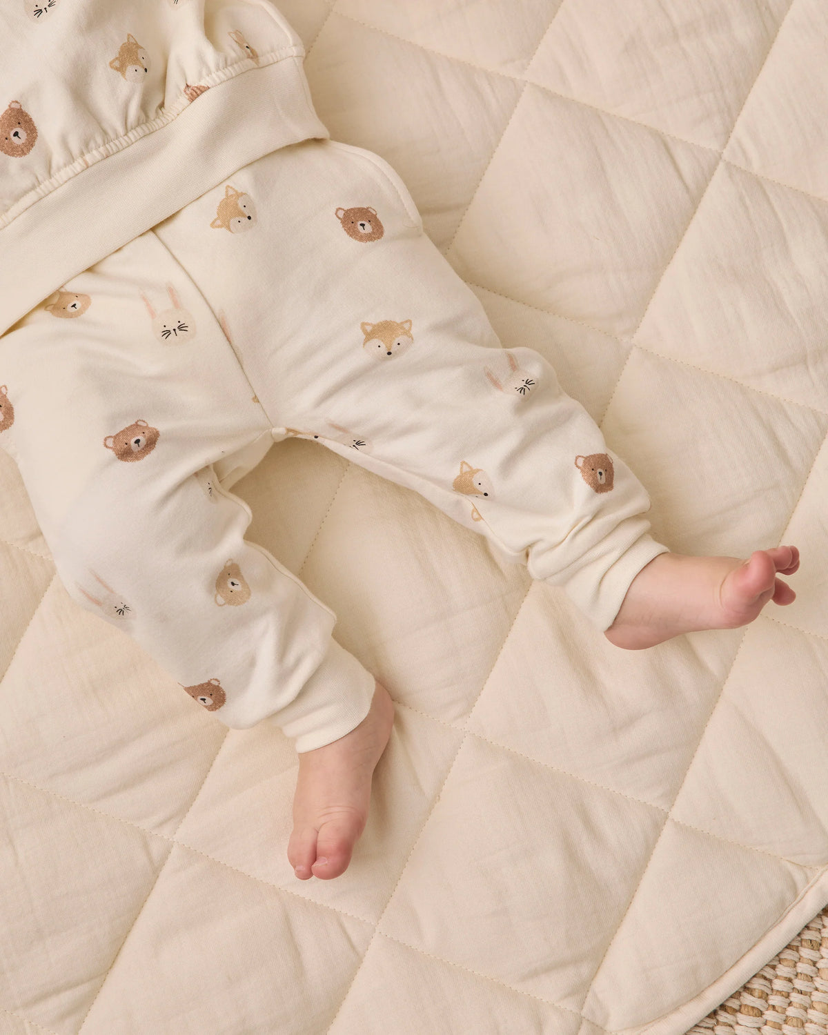 A baby lies on a cream-colored quilt, wearing Quincy Mae Baby Boys’ and Boys’ Relaxed Fleece Sweatpant featuring a cozy bear face pattern. With only the legs and lower torso visible, these sweatpants are an ideal choice for everyday comfort.