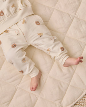 A baby lies on a cream-colored quilt, wearing Quincy Mae Baby Boys’ and Boys’ Relaxed Fleece Sweatpant featuring a cozy bear face pattern. With only the legs and lower torso visible, these sweatpants are an ideal choice for everyday comfort.