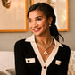 A woman in a black and cream blouse with gold jewelry and Berry Drop Stud Earrings by Juliska Capucine De Wulf smiles on a light sofa, subtle shimmer on her face, with framed art in the background.