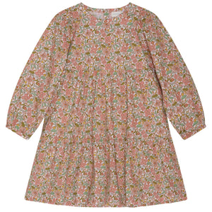 The Girls’ Puff Dress by Serendipity is a long-sleeved, knee-length organic cotton dress adorned with a hand-drawn flower print in pink, green, and white. It features a round neckline, subtle puff sleeves, and a loose, tiered silhouette.