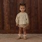A young child stands indoors on a rug, wearing the Rylee & Cru Baby Girls’ Quincy Set—a light patterned long-sleeve top and matching bloomer—along with ruffle socks and tan shoes, in front of a wooden wall.