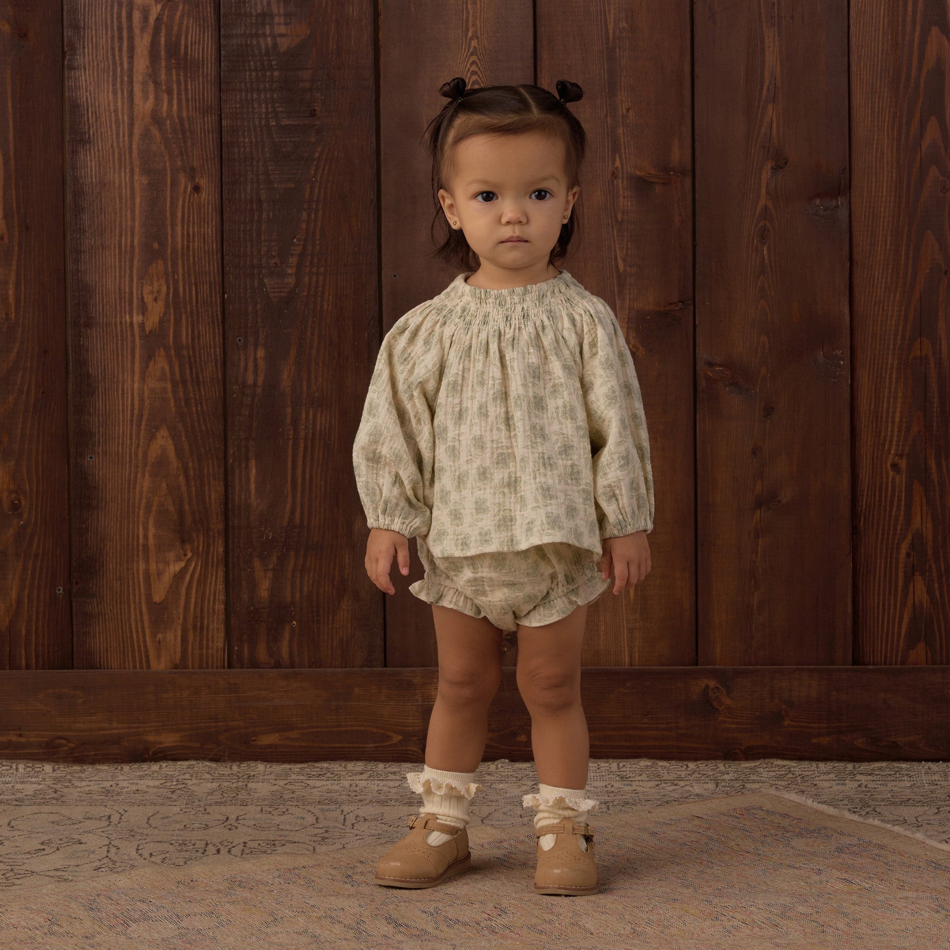 A young child stands indoors on a rug, wearing the Rylee & Cru Baby Girls’ Quincy Set—a light patterned long-sleeve top and matching bloomer—along with ruffle socks and tan shoes, in front of a wooden wall.
