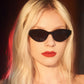 A person with long blonde hair wears Thierry Lasry Acidity sunglasses, handmade in France, in a stylish cat-eye design that offers UV protection. They pair the look with bright red lipstick against a softly lit background featuring red and warm tones.