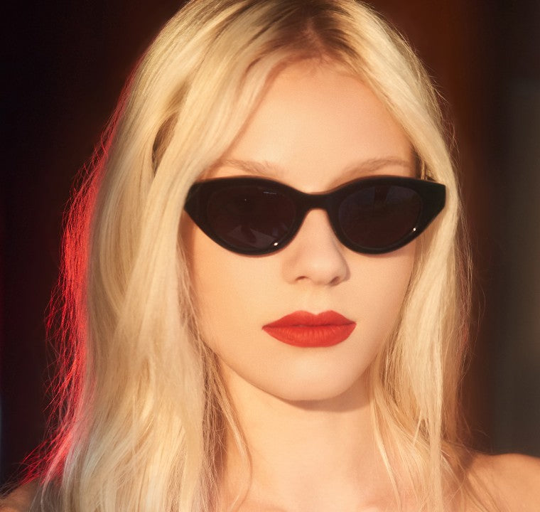 A person with long blonde hair wears Thierry Lasry Acidity sunglasses, handmade in France, in a stylish cat-eye design that offers UV protection. They pair the look with bright red lipstick against a softly lit background featuring red and warm tones.