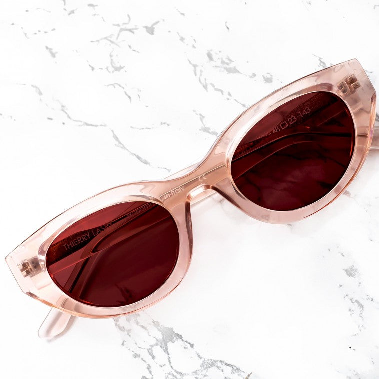 The Thierry Lasry Acidity Sunglasses, featuring oval lenses and clear pink frames, rest on a white marble surface, offering UV protection. Handmade in France by the renowned brand Thierry Lasry, these chic shades exude elegance and style.