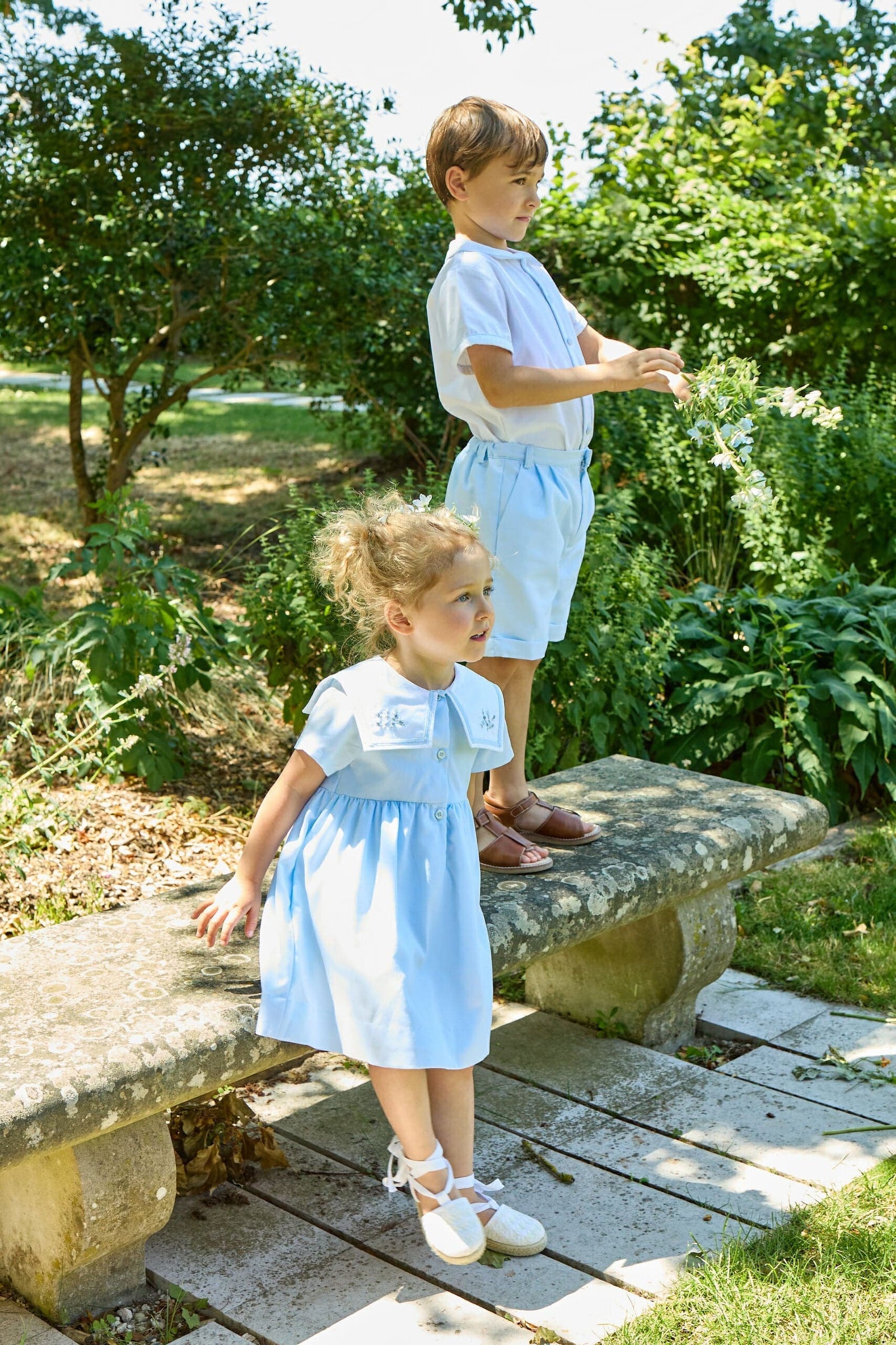 Two young boys wear Kidiwi's Baby Boys' and Boys' Noa MC Shirt as they stand on and beside a stone bench in a sunlit garden surrounded by greenery.