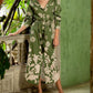 A woman stands on a balcony dressed in an Andres Otalora Armenia Dress, showcasing a green and white print long-sleeve design with a matching belt. Made from 100% cotton, she complements the outfit with brown sandals. The surroundings are lush with greenery and a green wall.