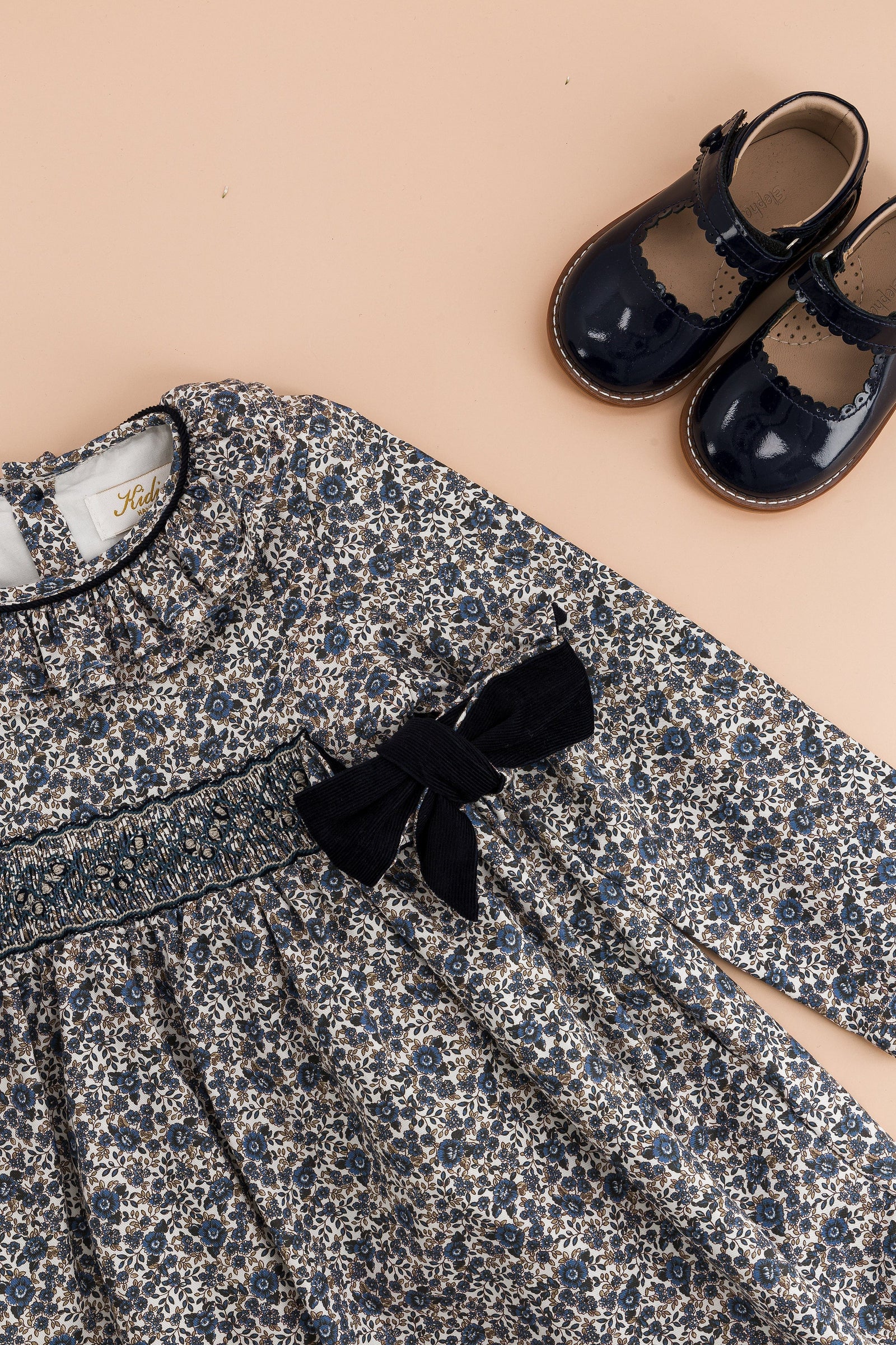 The Kidiwi Baby Girls' and Girls' Adriana Corduroy Bows Smocked Dress is displayed next to shiny navy blue Mary Jane shoes, all set against a beige background.