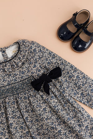 The Kidiwi Baby Girls' and Girls' Adriana Corduroy Bows Smocked Dress is displayed next to shiny navy blue Mary Jane shoes, all set against a beige background.