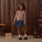 A young girl stands indoors on a rug in front of a wooden wall, wearing a pink cardigan, denim skirt, white socks, and black shoes, with her curly hair adorned by the Rylee & Cru Girls’ Bow for a charming touch.