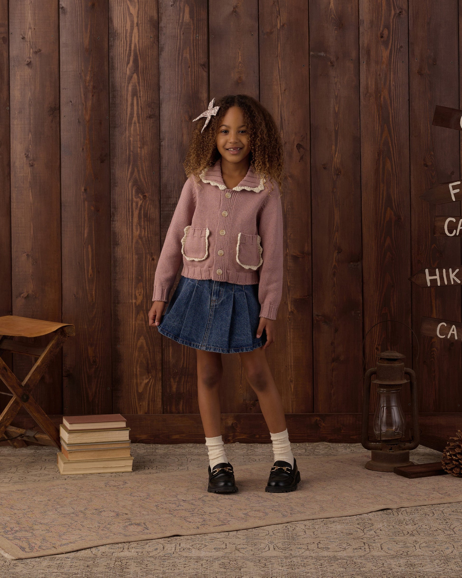 A young girl stands indoors on a rug in front of a wooden wall, wearing a pink cardigan, denim skirt, white socks, and black shoes, with her curly hair adorned by the Rylee & Cru Girls’ Bow for a charming touch.