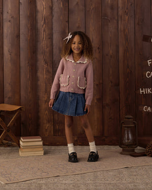 A young girl stands indoors on a rug in front of a wooden wall, wearing a pink cardigan, denim skirt, white socks, and black shoes, with her curly hair adorned by the Rylee & Cru Girls’ Bow for a charming touch.