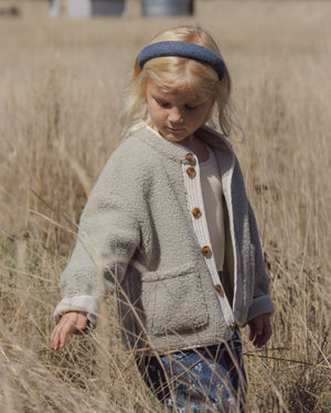 A young blonde-haired child with a blue headband stands in a dry field, gently touching tall grass while wearing the Rylee & Cru Baby Girls’ and Girls’ Chalet Jacket, featuring a cozy sherpa design and quilted button placket.
