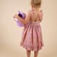 A young girl wears the Early Sunday Girls’ Alena Dress in a floral purple print as she stands facing away, holding a purple watering can against a plain beige background.