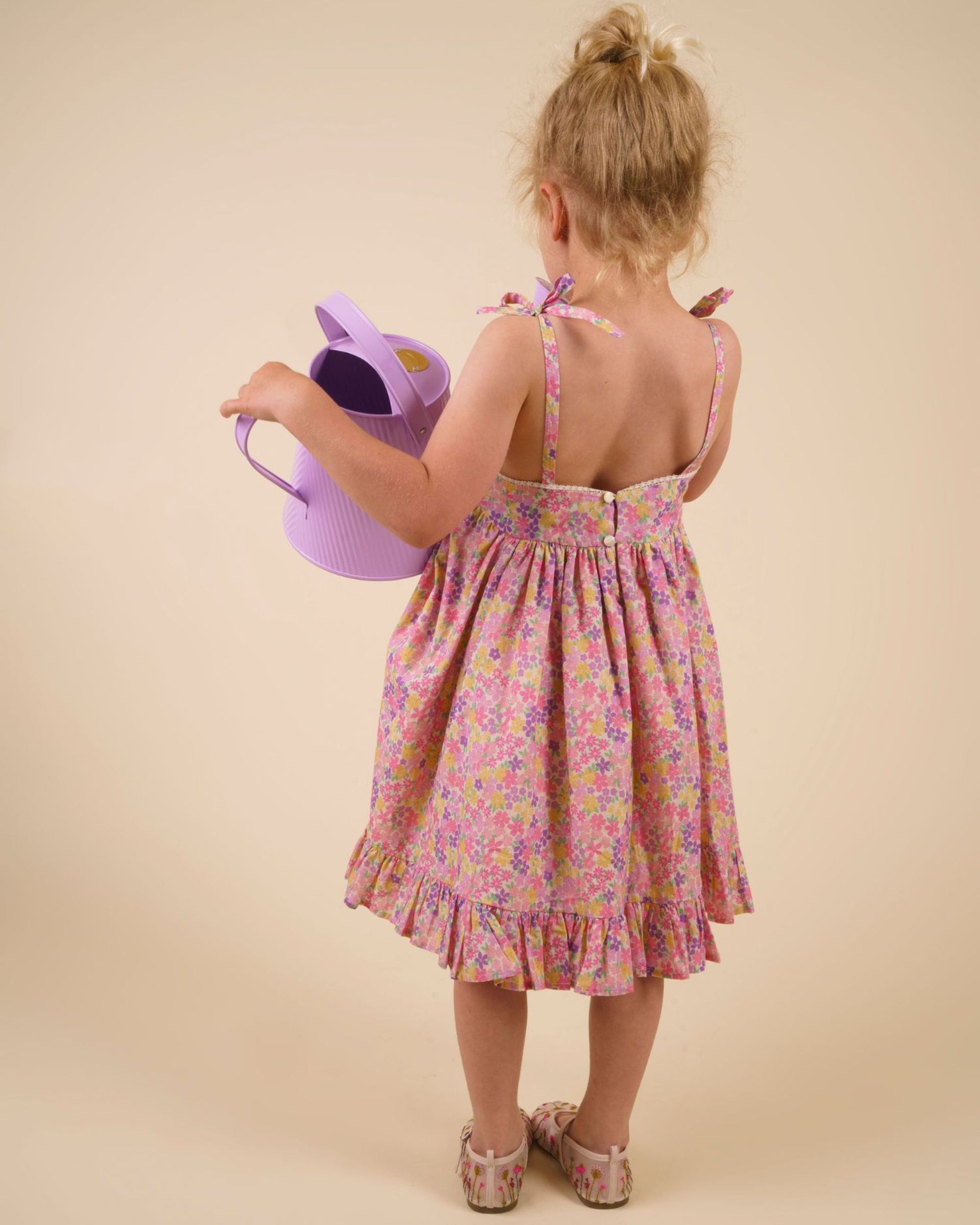 A young girl wears the Early Sunday Girls’ Alena Dress in a floral purple print as she stands facing away, holding a purple watering can against a plain beige background.