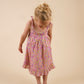 A young child with blonde hair wearing the Early Sunday Girls’ Alena Dress and sandals stands against a plain beige background, looking down at the floor.
