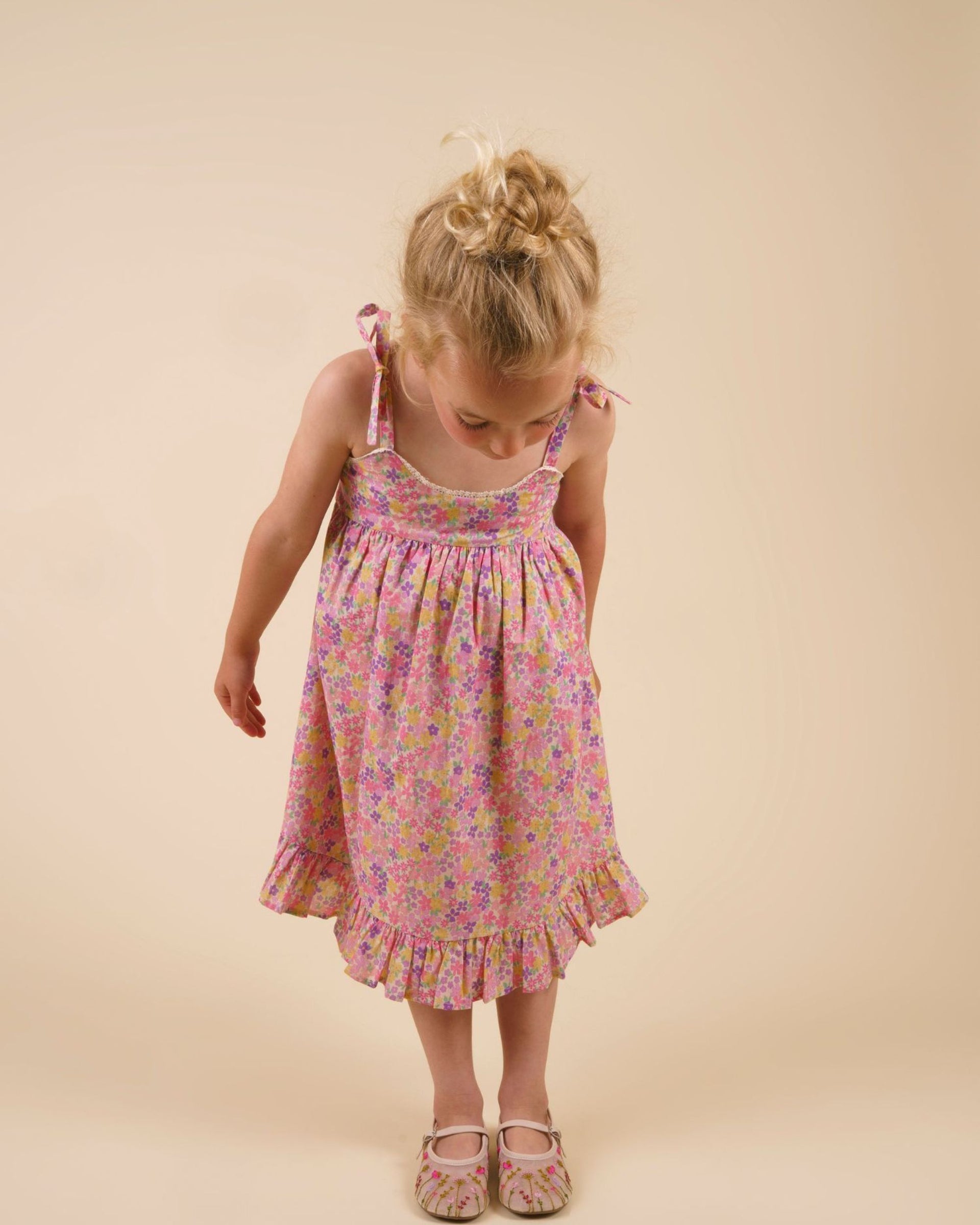 A young child with blonde hair wearing the Early Sunday Girls’ Alena Dress and sandals stands against a plain beige background, looking down at the floor.