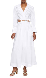 A person is wearing the Simkhai Signature Alex Dress, a long white dress with stylish cutouts at the waist and long sleeves, featuring a wrap accent, paired with light-colored heeled sandals from the brand Simkhai.