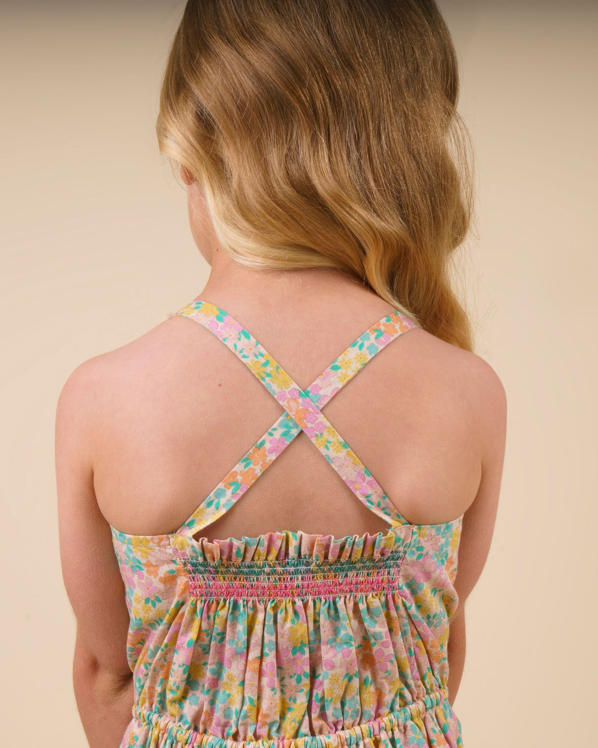 A young girl with light brown hair is shown from the back, wearing the Early Sunday Girls’ Alisson Dress, a floral mint dress featuring crisscross straps and gathered elastic at the top.