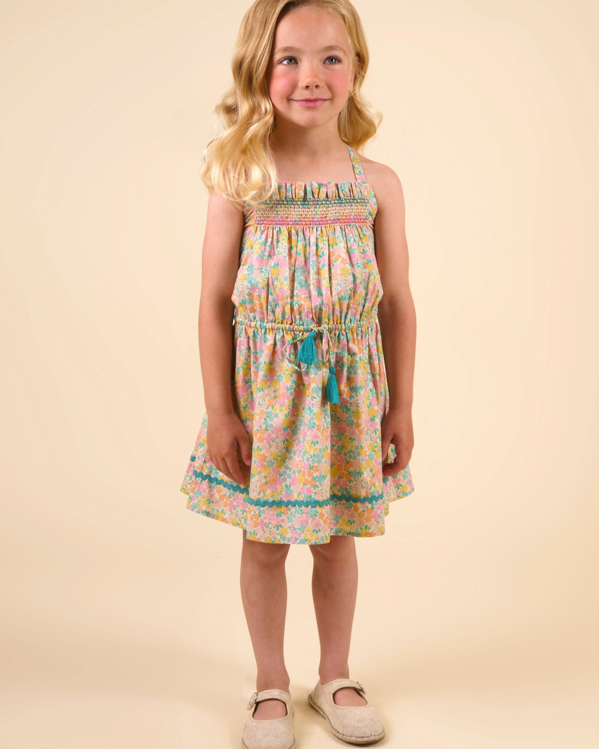 A young girl with blonde hair stands facing forward in the Early Sunday Girls’ Alisson Dress, crafted from organic cotton with a mint floral pattern, paired with beige shoes against a plain, light beige background.