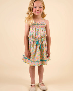 A young girl with blonde hair stands facing forward in the Early Sunday Girls’ Alisson Dress, crafted from organic cotton with a mint floral pattern, paired with beige shoes against a plain, light beige background.