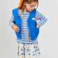 A young girl wears the Arsene & Les Pipelettes Girls' Aloe Skirt featuring a gold elasticated waistband, paired with a striped long-sleeve shirt, a blue vest, and an orange scarf tied in her hair. She looks down with one hand tucked in her vest pocket against a simple background.